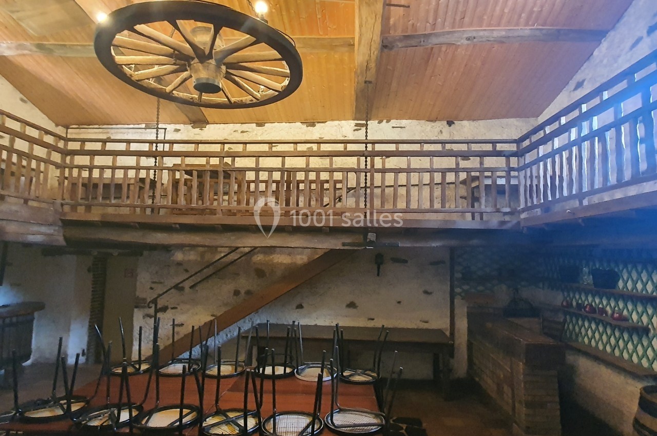 Salle rustique avec plafond en bois, mezzanine en balustrade, grande table dressée et chaises retournées.