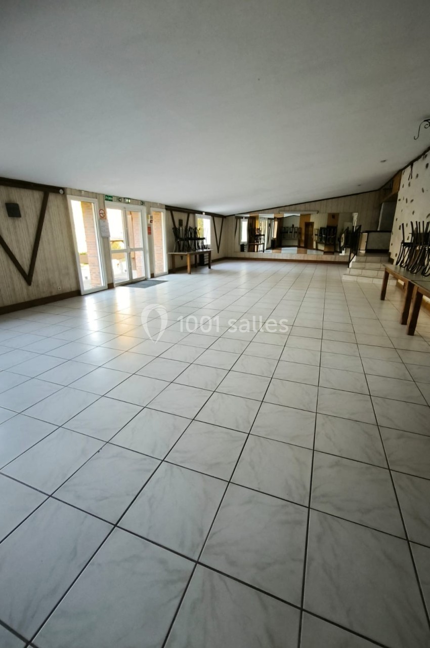 Salle vide avec sol carrelé blanc, murs décorés de bois, miroirs et portes vitrées donnant sur l'extérieur.