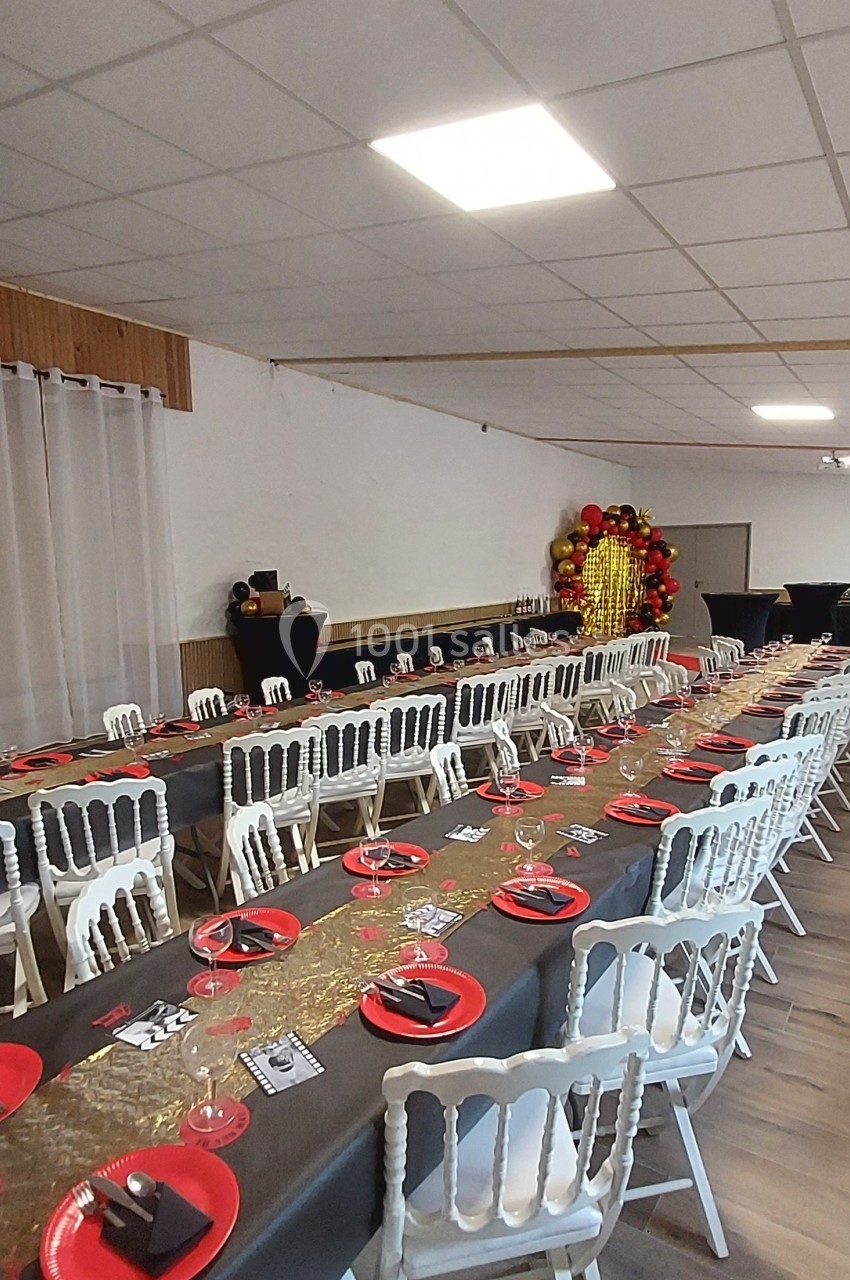 Salle décorée pour un événement avec longues tables dressées, nappes noires, assiettes rouges et décoration dorée en fond.