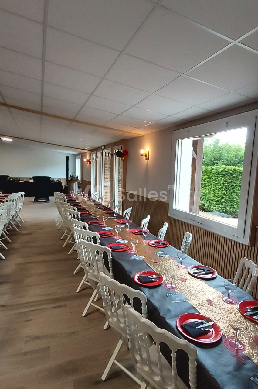 Salle décorée avec une longue table dressée, nappes noires et dorées, assiettes rouges, chaises blanches, grandes fenêtres.