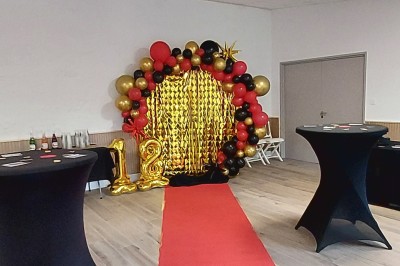 Décoration festive avec arche de ballons rouges, noirs et dorés, rideau scintillant et tapis rouge dans une salle.