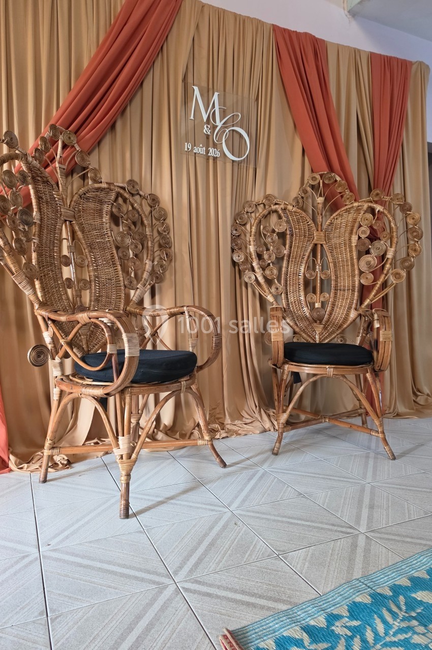 Deux fauteuils en rotin ornés, disposés devant un rideau beige et orange avec des initiales et une date en arrière-plan.