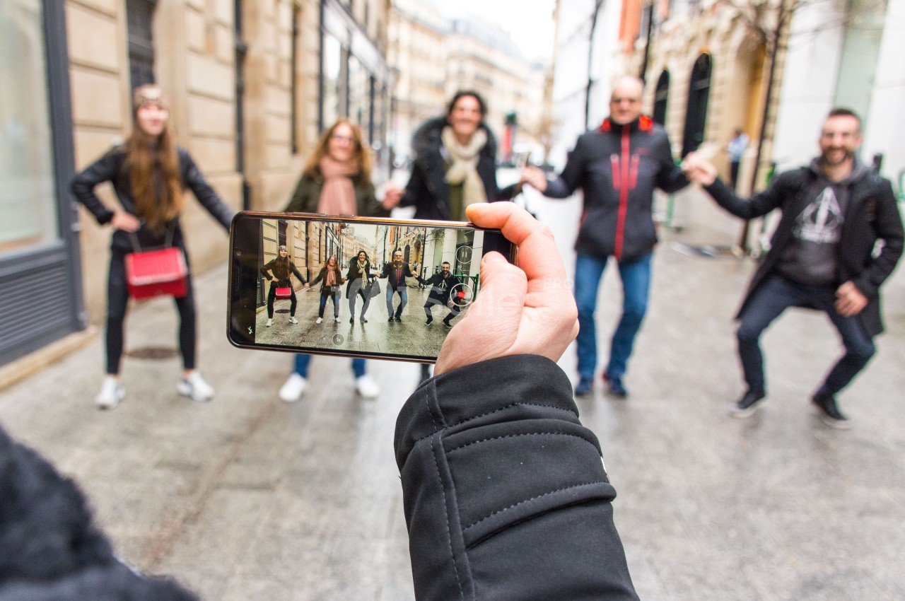 Une personne filme un groupe de cinq personnes dansant dans une rue urbaine avec un smartphone.