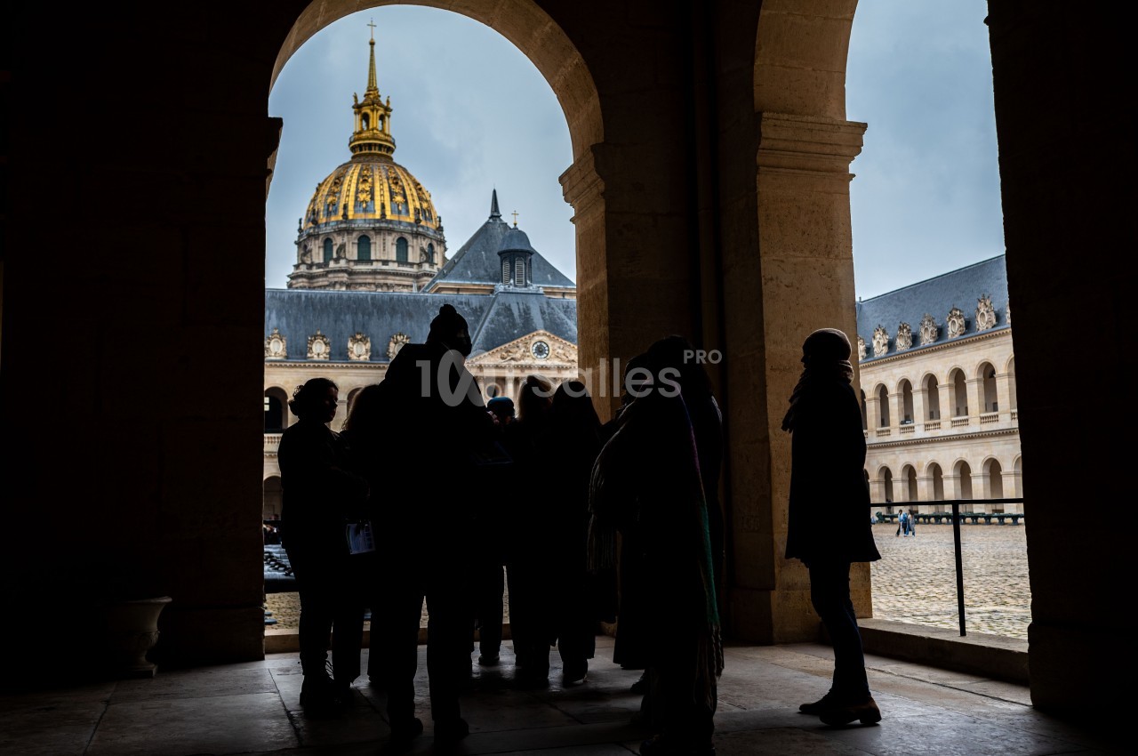 Groupe de personnes en silhouette sous une arcade, avec le dôme doré des Invalides visible en arrière-plan.
