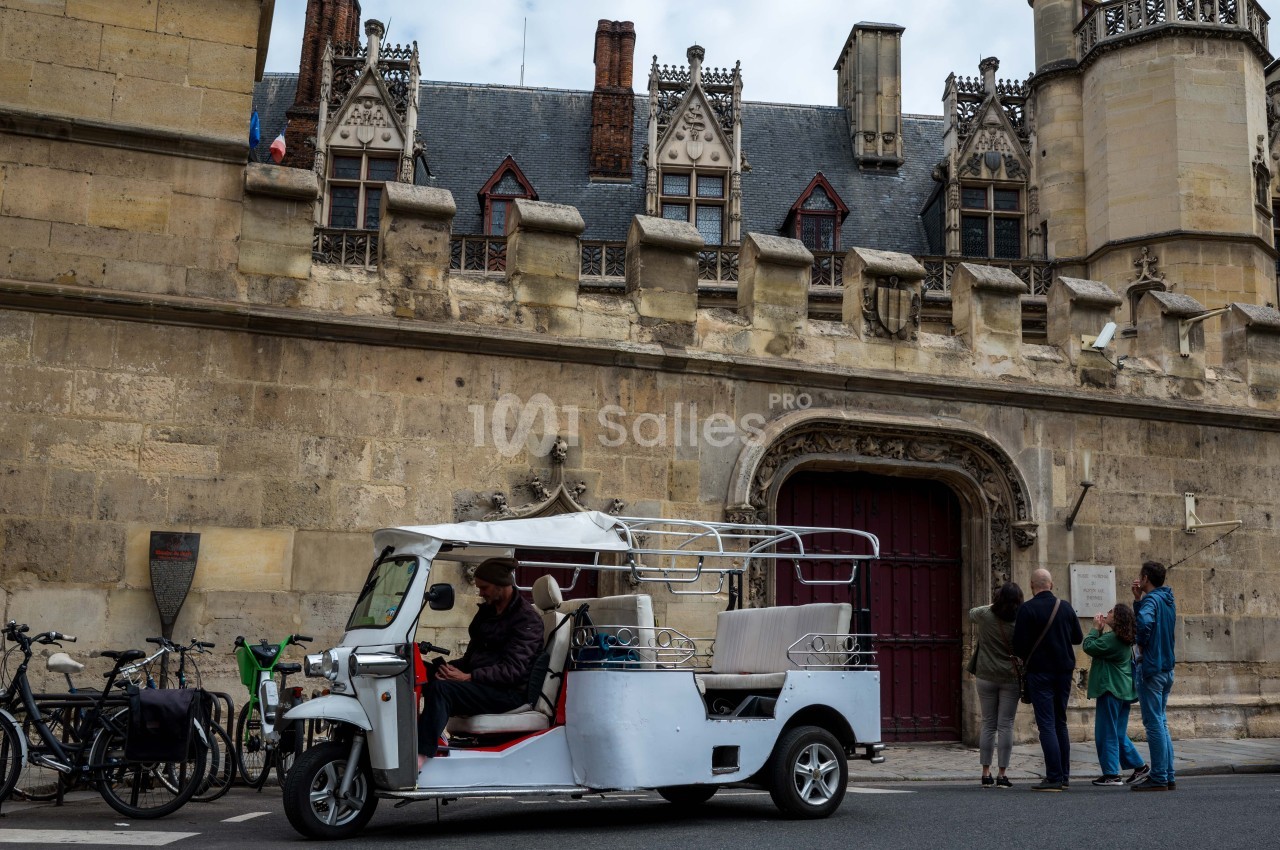 Un tuk-tuk blanc stationné devant un bâtiment ancien en pierre avec des fenêtres gothiques et des passants observant.