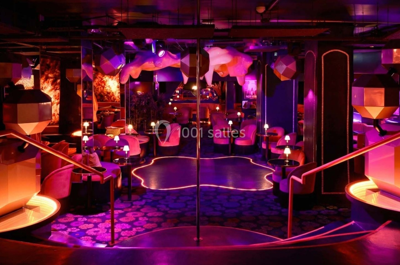 Salle de club avec éclairage néon rose et violet, tables rondes, fauteuils, et décorations géométriques modernes.