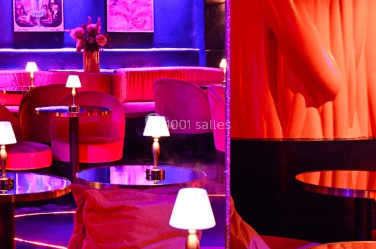 Salle de bar avec éclairage tamisé, tables rondes, petites lampes et fauteuils rouges dans une ambiance chaleureuse.