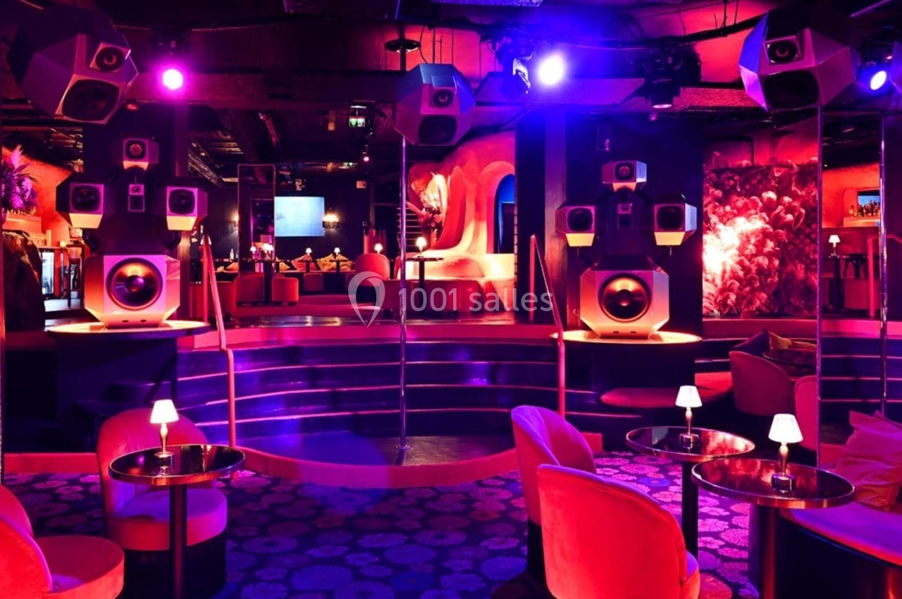 Salle de club colorée avec éclairage violet, sièges confortables, tables basses et enceintes disposées autour d'une scène.