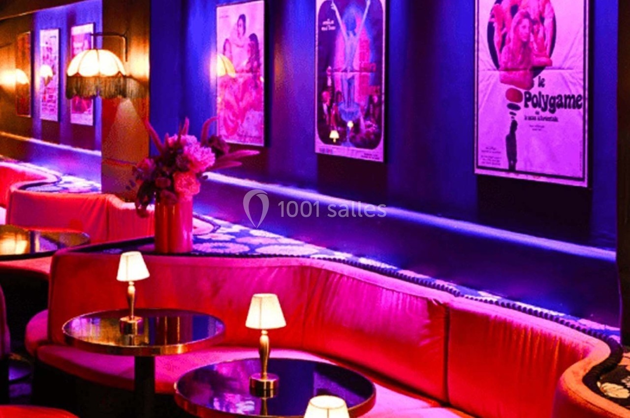 Banquettes rouges, petites lampes et affiches rétro éclairées dans un bar à l'ambiance tamisée et colorée.