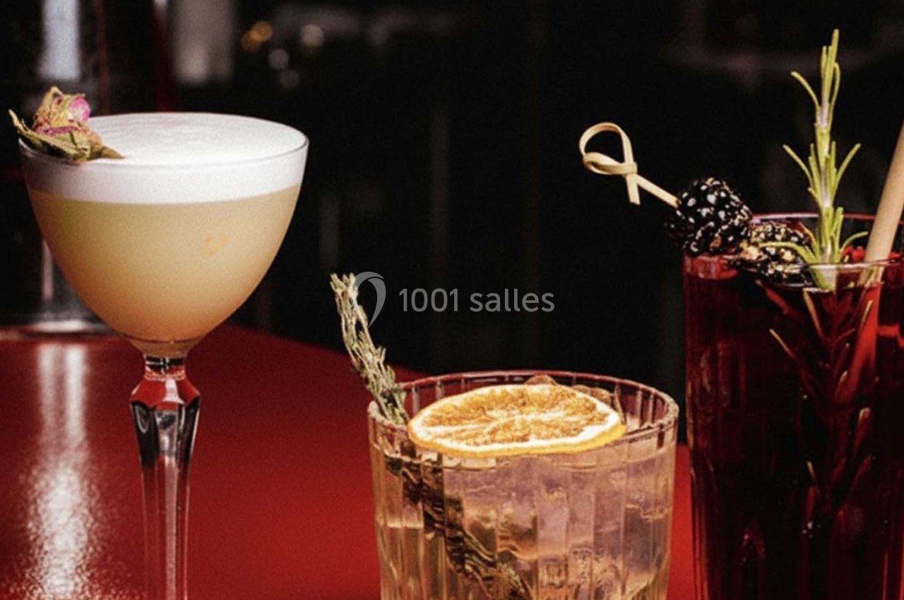 Trois cocktails variés sur une table rouge, décorés avec des herbes, fruits séchés et garnitures originales.