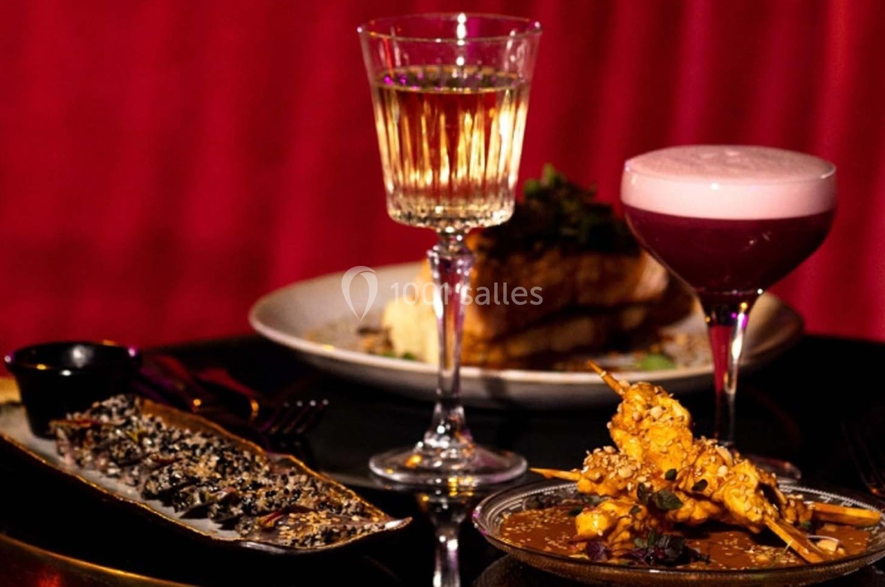 Assiette de brochettes et plat de poisson avec deux cocktails sur une table, fond rouge tamisé.