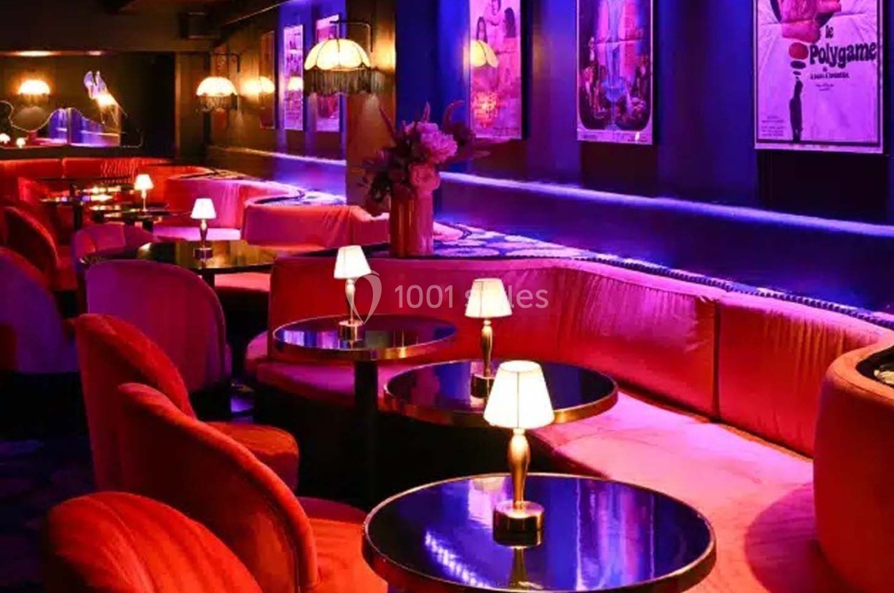 Salle de bar lounge avec banquettes roses, tables rondes noires, petites lampes allumées et affiches rétro au mur.