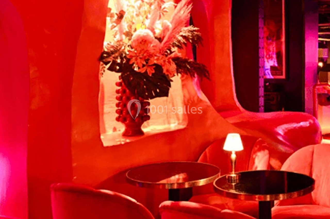 Décor intérieur avec fauteuils rouges, tables noires et un vase fleuri éclairé par une lumière tamisée.