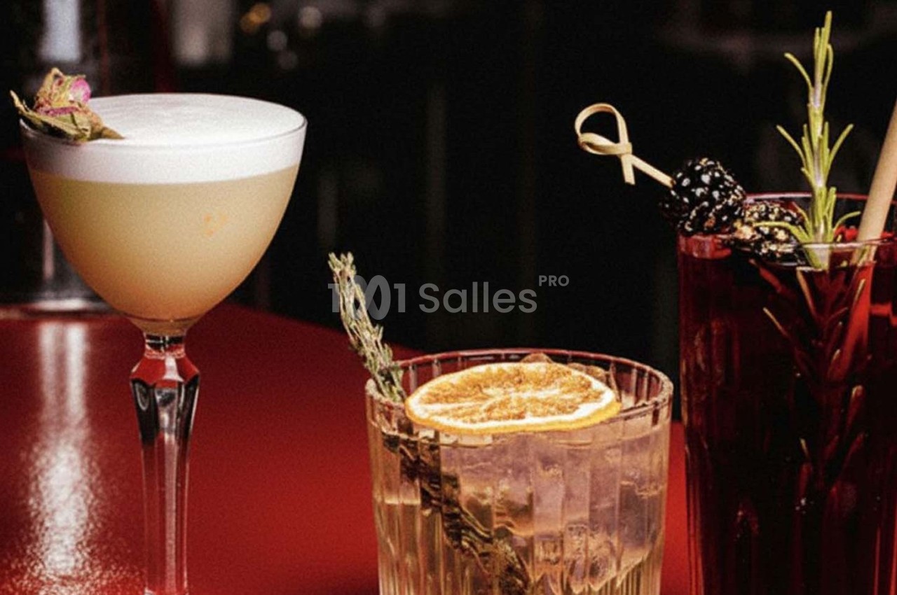 Trois cocktails variés sur une table rouge, décorés avec des fruits, herbes et garnitures élégantes.
