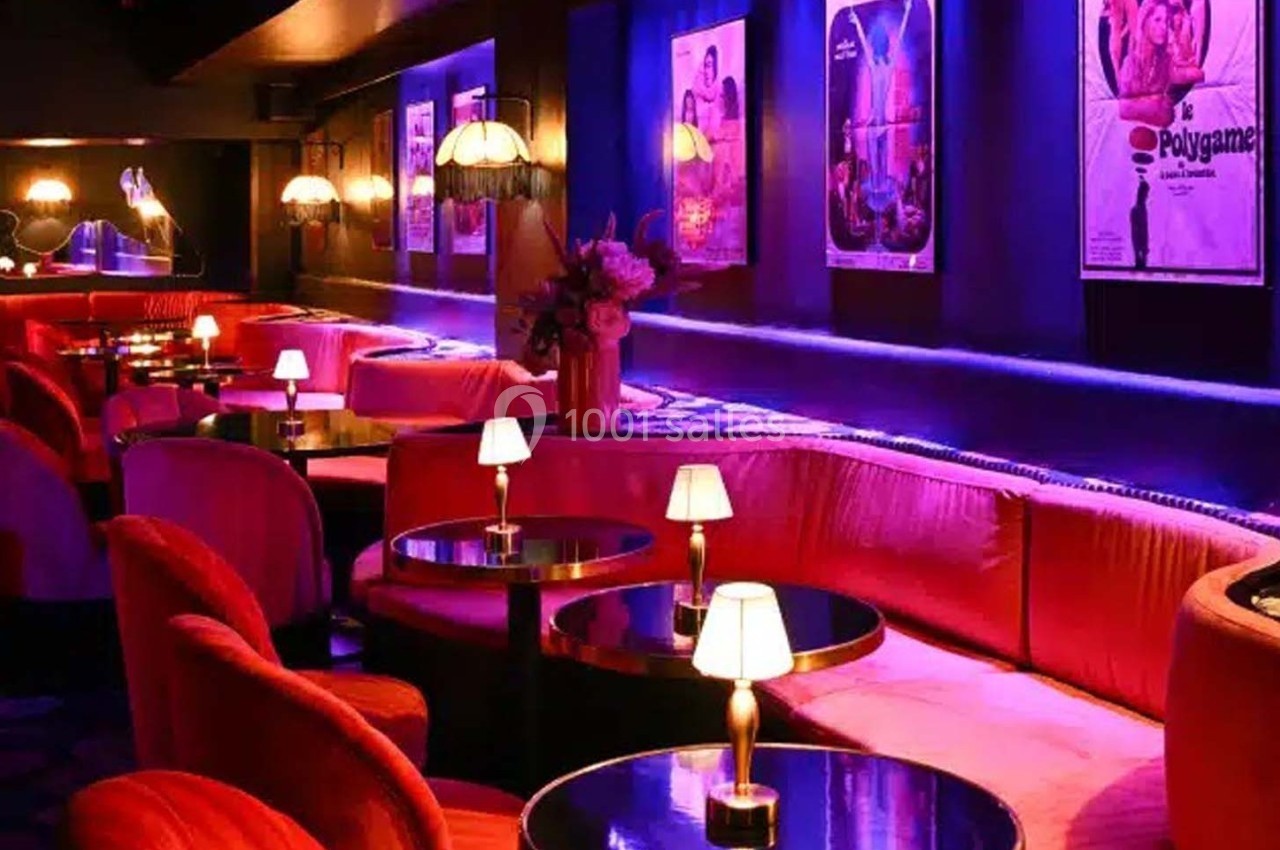 Salle de bar avec banquettes rouges, petites tables rondes et lampes tamisées, décorée d'affiches rétro au mur.