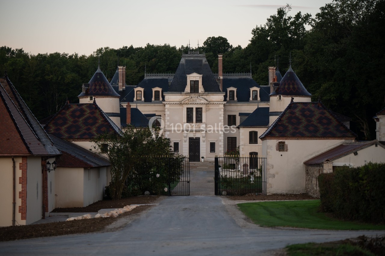 Façade d'un château entouré de dépendances, avec une allée centrale menant à une grille en fer forgé.