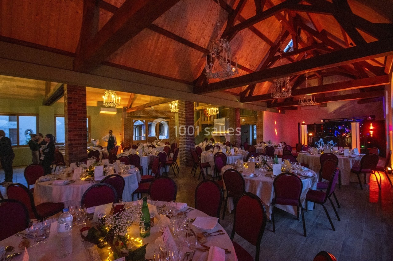 Salle de réception avec tables rondes dressées, éclairage chaleureux et poutres apparentes en bois.