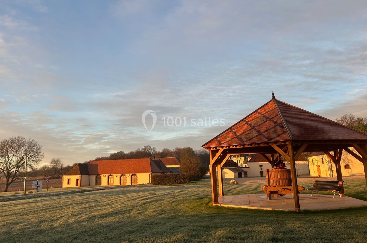 Abri en bois avec un pressoir, pelouse givrée et bâtiments en arrière-plan sous un ciel dégagé au lever du soleil.