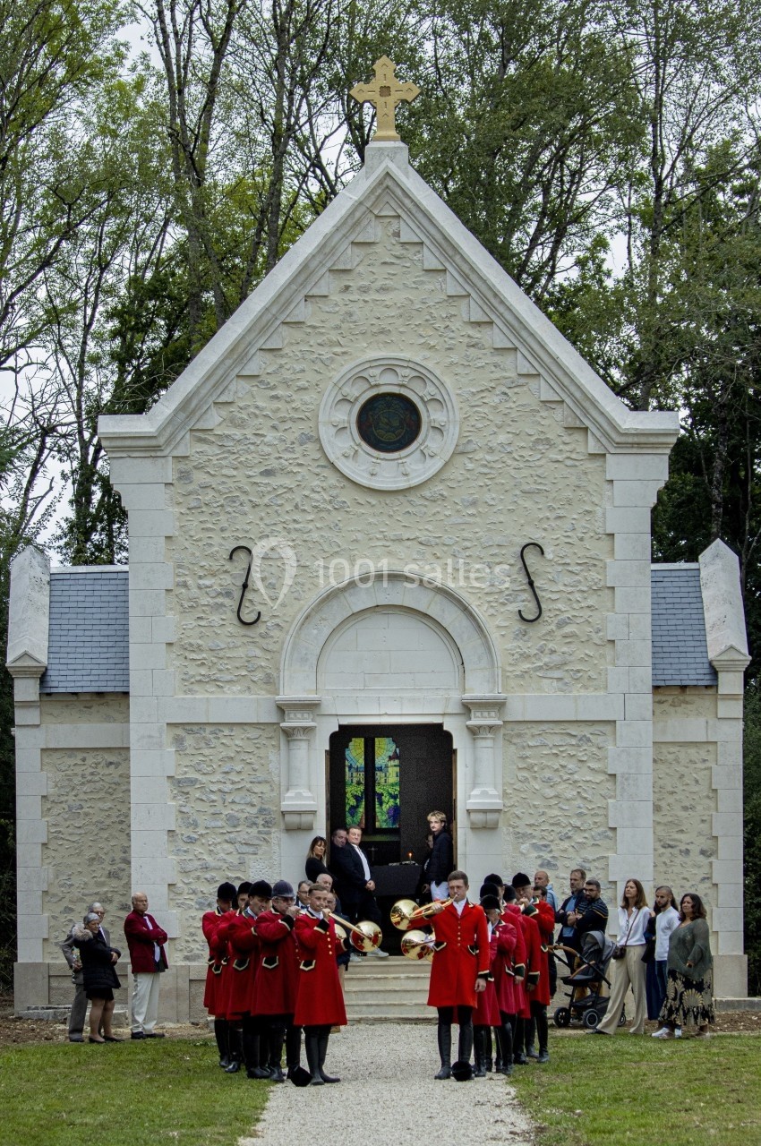 Un groupe de personnes en uniforme rouge et noir se tient devant une chapelle en pierre entourée d'arbres.