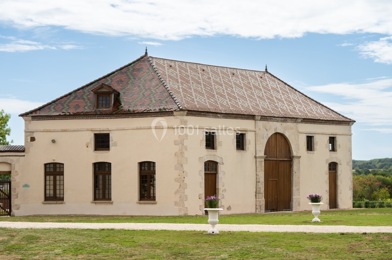 Bâtiment ancien avec toit en tuiles, grandes portes en bois et fenêtres, entouré de pelouse et de jardinières fleuries.