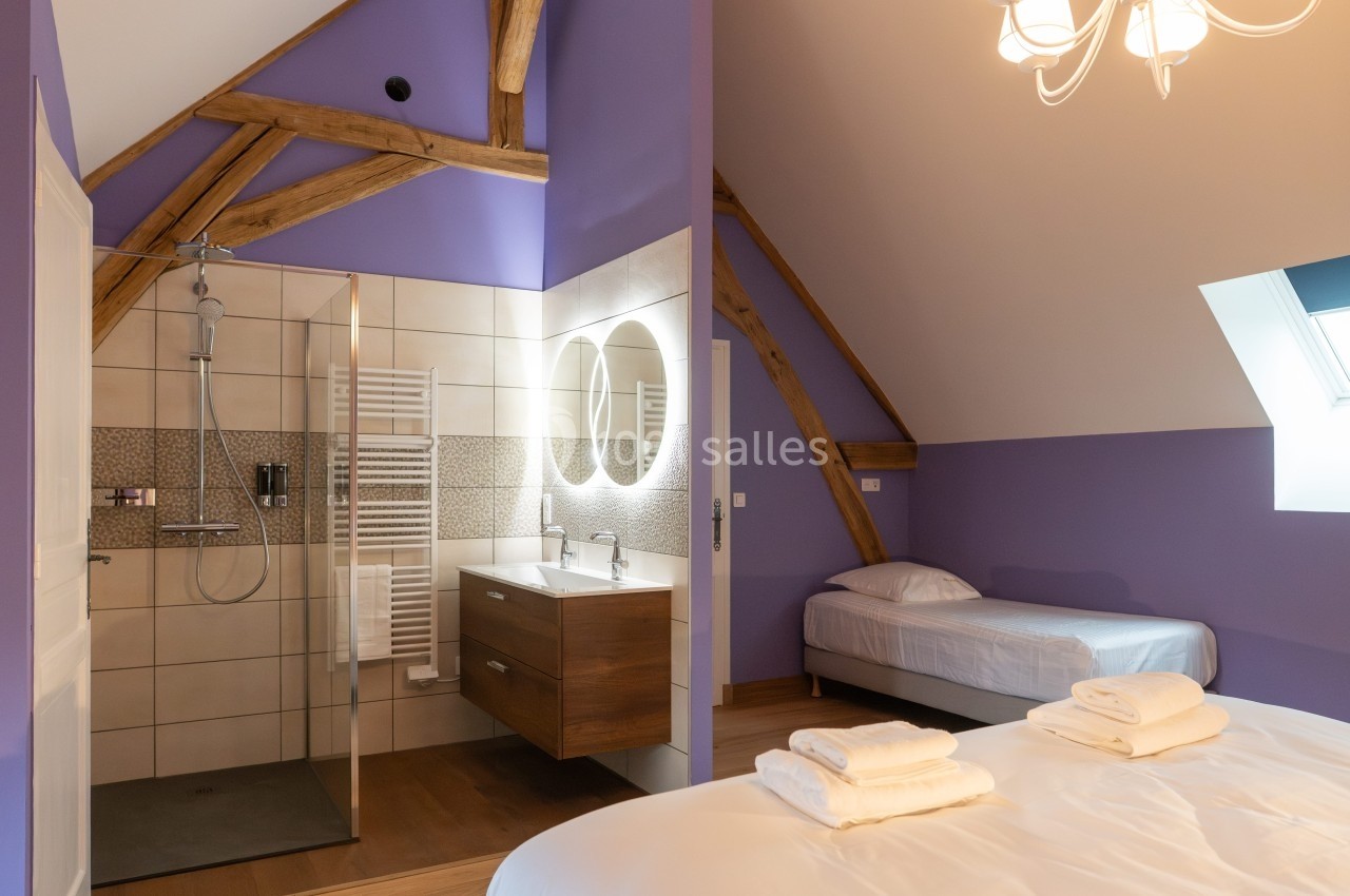 Salle de bain avec douche à l'italienne, lavabo double, murs violets, lit simple et lit double sous un plafond mansardé.