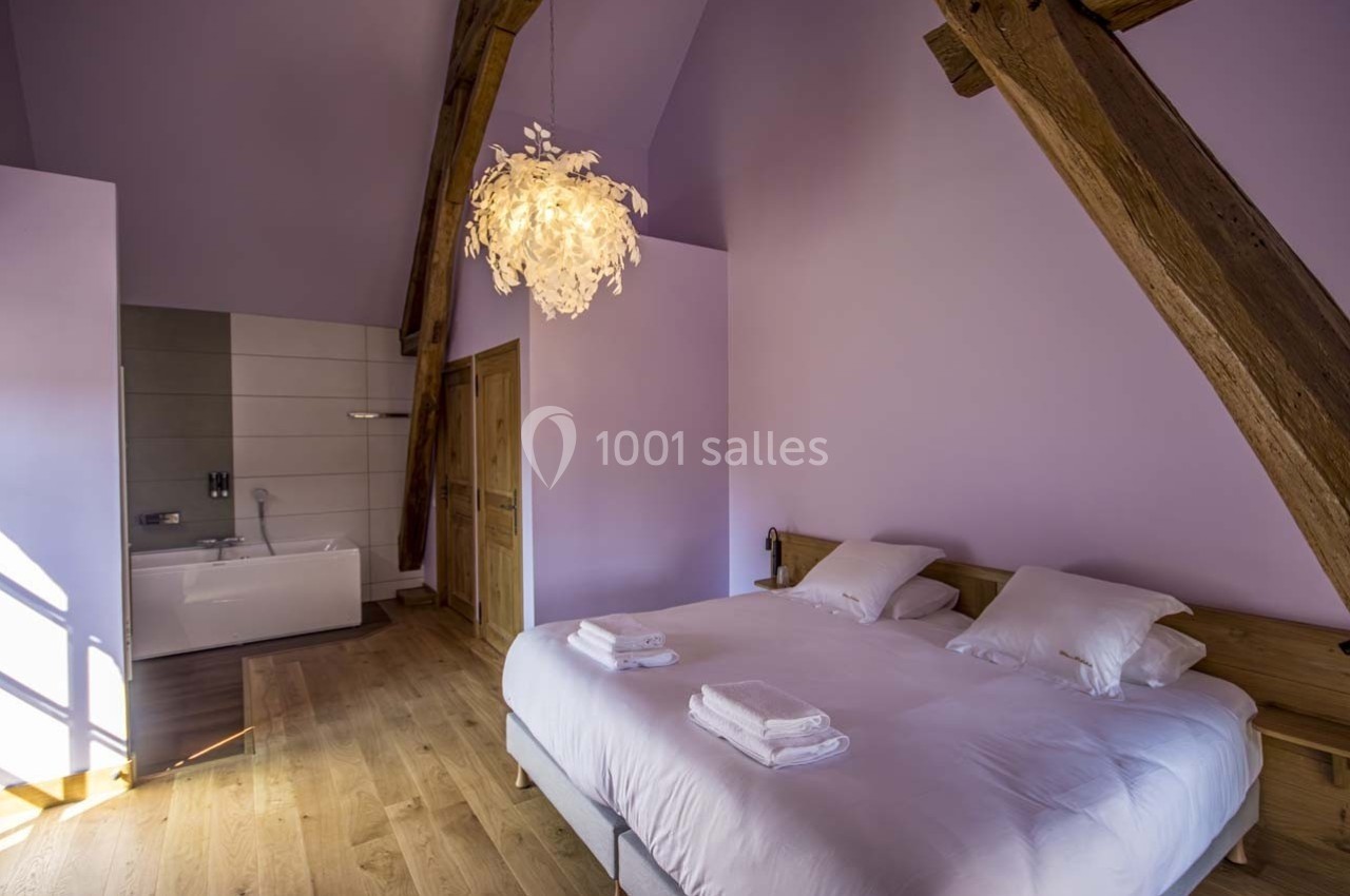 Chambre lumineuse avec lit double, poutres apparentes, parquet en bois et salle de bain attenante avec baignoire.