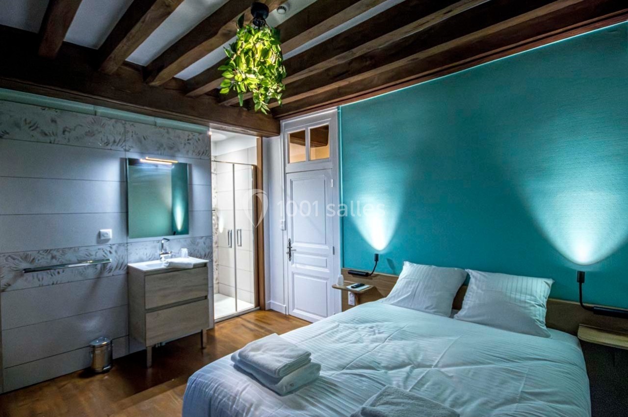 Chambre lumineuse avec lit double, salle de bain attenante, murs turquoise et poutres apparentes en bois.