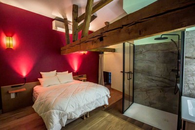 Chambre avec lit double, poutres apparentes, linge de lit blanc, et salle de bain attenante visible par une porte ouverte.