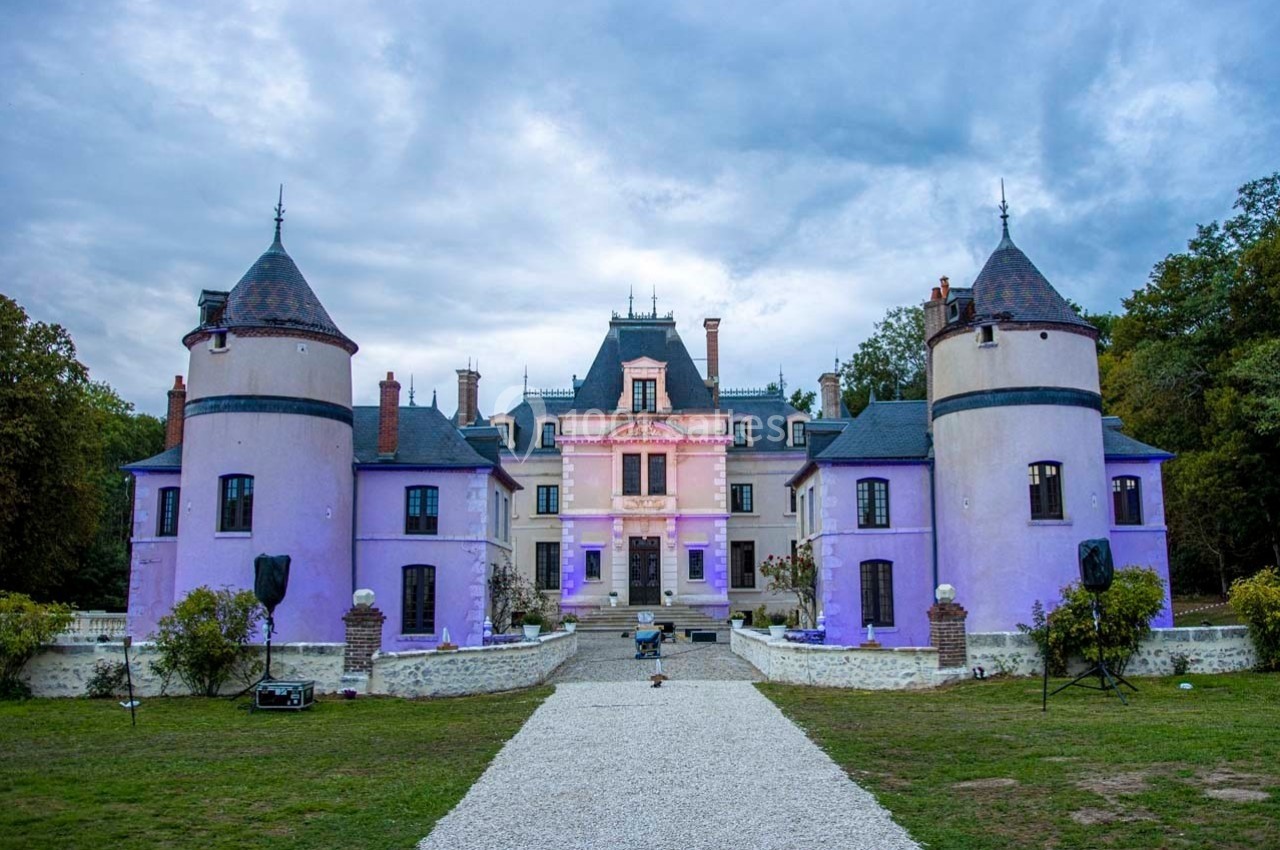 Façade d'un château éclairé de lumières violettes, entouré de tours et d'un jardin avec une allée centrale en gravier.