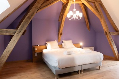 Chambre avec lit double, poutres apparentes, linge de lit blanc, et salle de bain attenante visible par une porte ouverte.