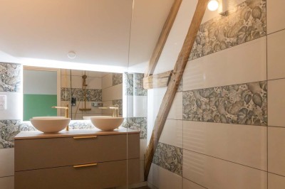 Chambre avec lit double, poutres apparentes, linge de lit blanc, et salle de bain attenante visible par une porte ouverte.