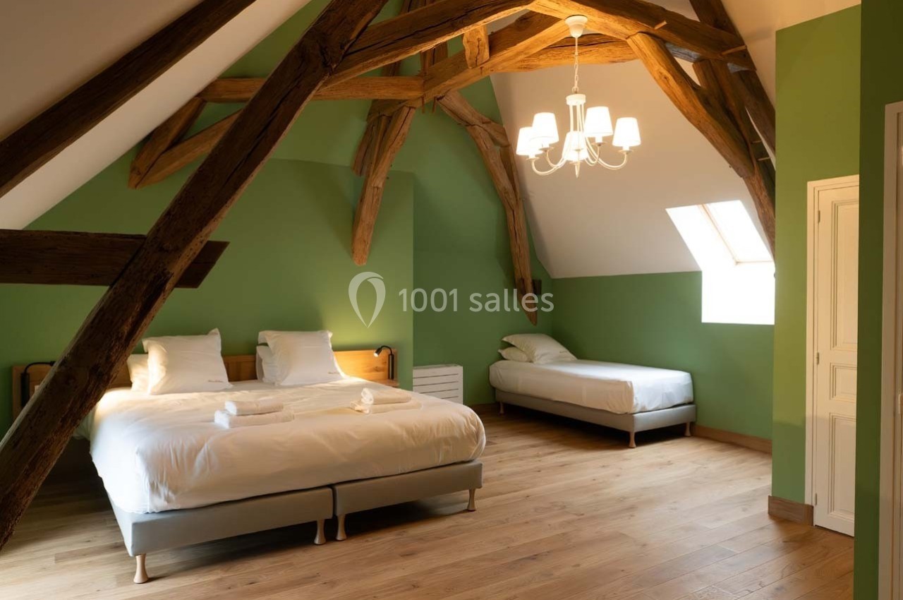 Chambre mansardée avec poutres apparentes, murs verts, deux lits, parquet en bois et lumière naturelle.