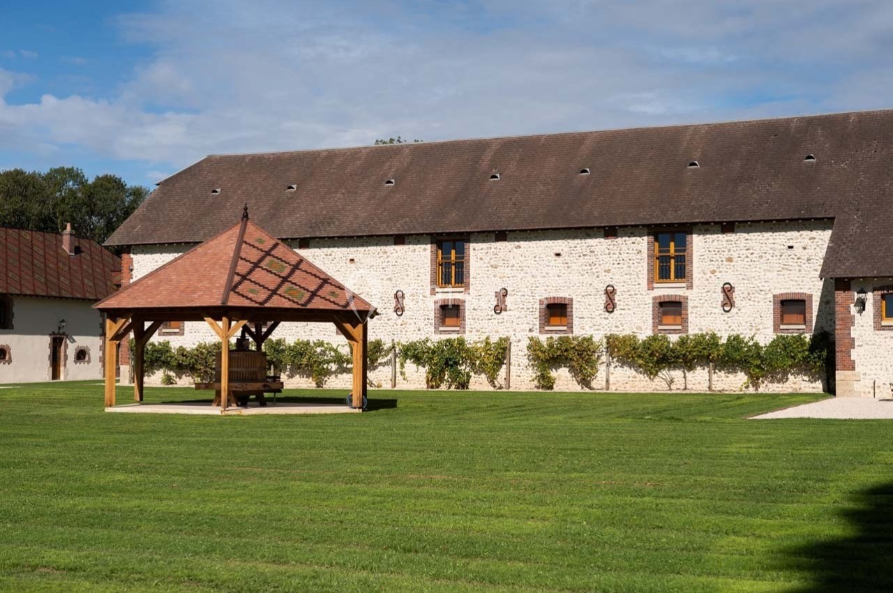 Bâtiment en pierre avec toit en ardoise, pergola en bois au premier plan et pelouse bien entretenue.