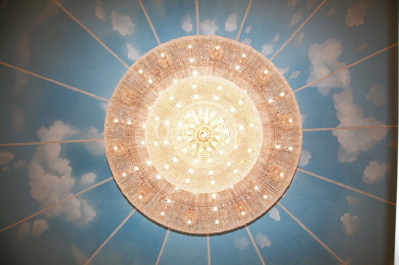 Lustre circulaire en cristal éclairé, suspendu sous un plafond peint avec des nuages sur un fond bleu.