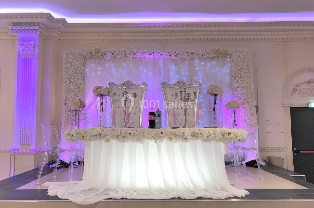 Deux chaises décorées sur une estrade ornée de fleurs blanches et de rideaux lumineux dans une salle de réception.