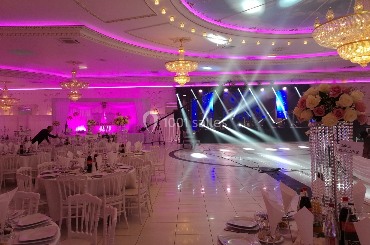Salle de réception élégante avec tables dressées, lustres scintillants et éclairage coloré sur une scène.