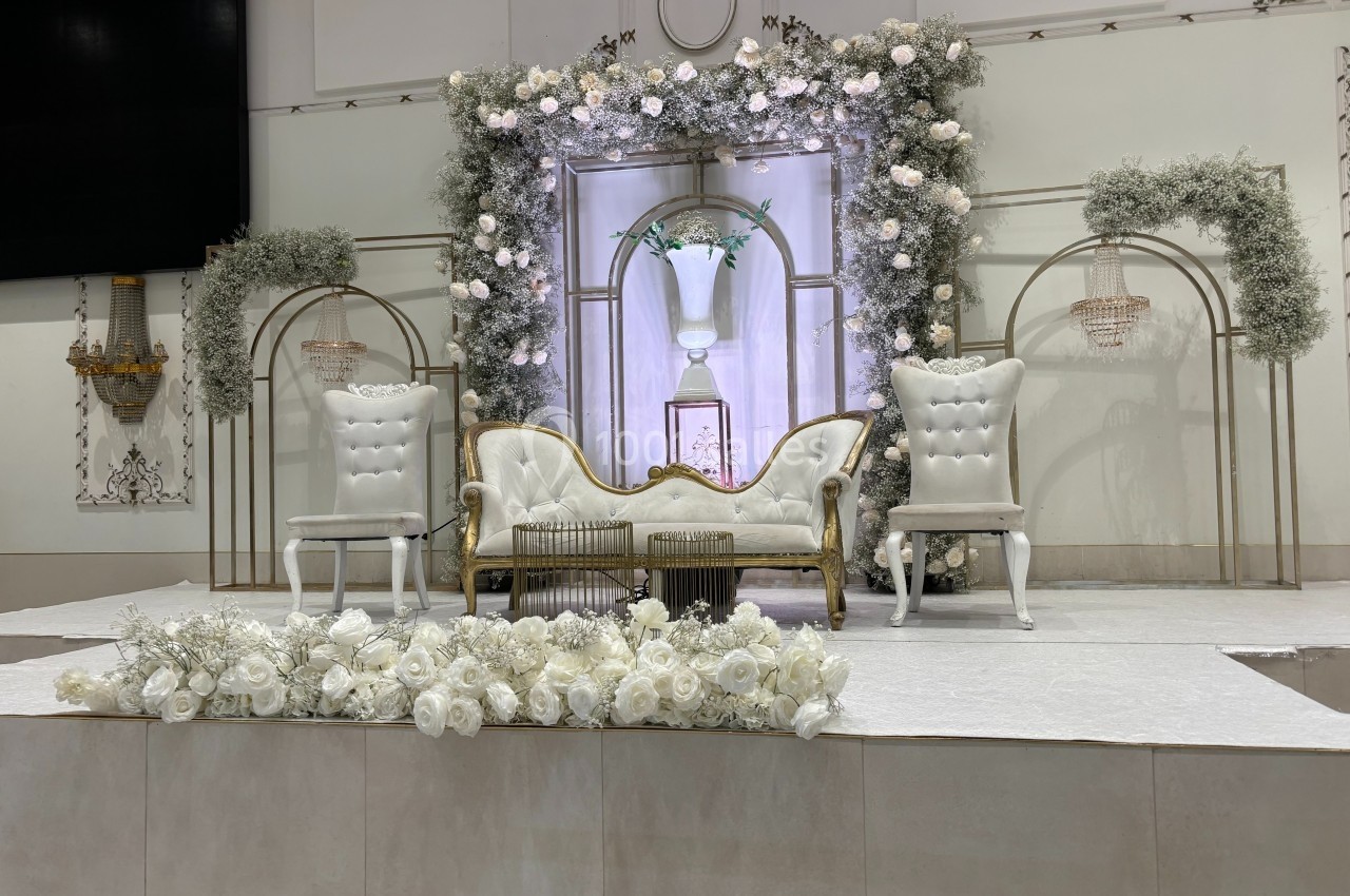 Décor élégant avec canapé doré, fauteuils blancs et arche ornée de fleurs blanches dans une salle de réception.