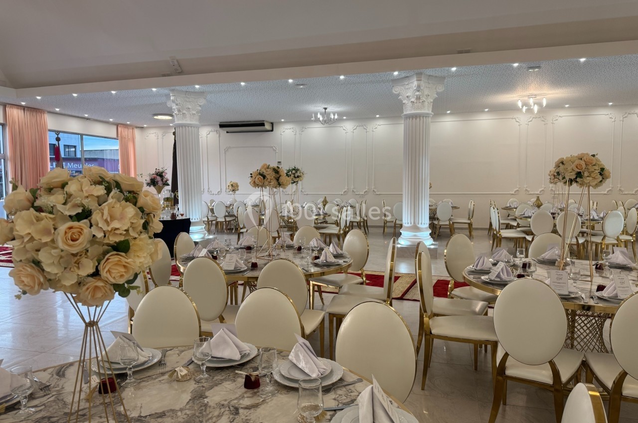 Salle de réception élégante avec tables rondes dressées, chaises blanches et décorations florales en tons crème.