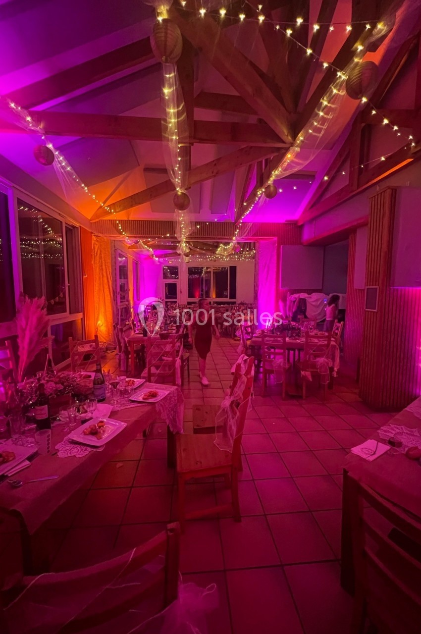Salle décorée pour un événement festif avec guirlandes lumineuses, tables dressées et éclairage rose et orange.