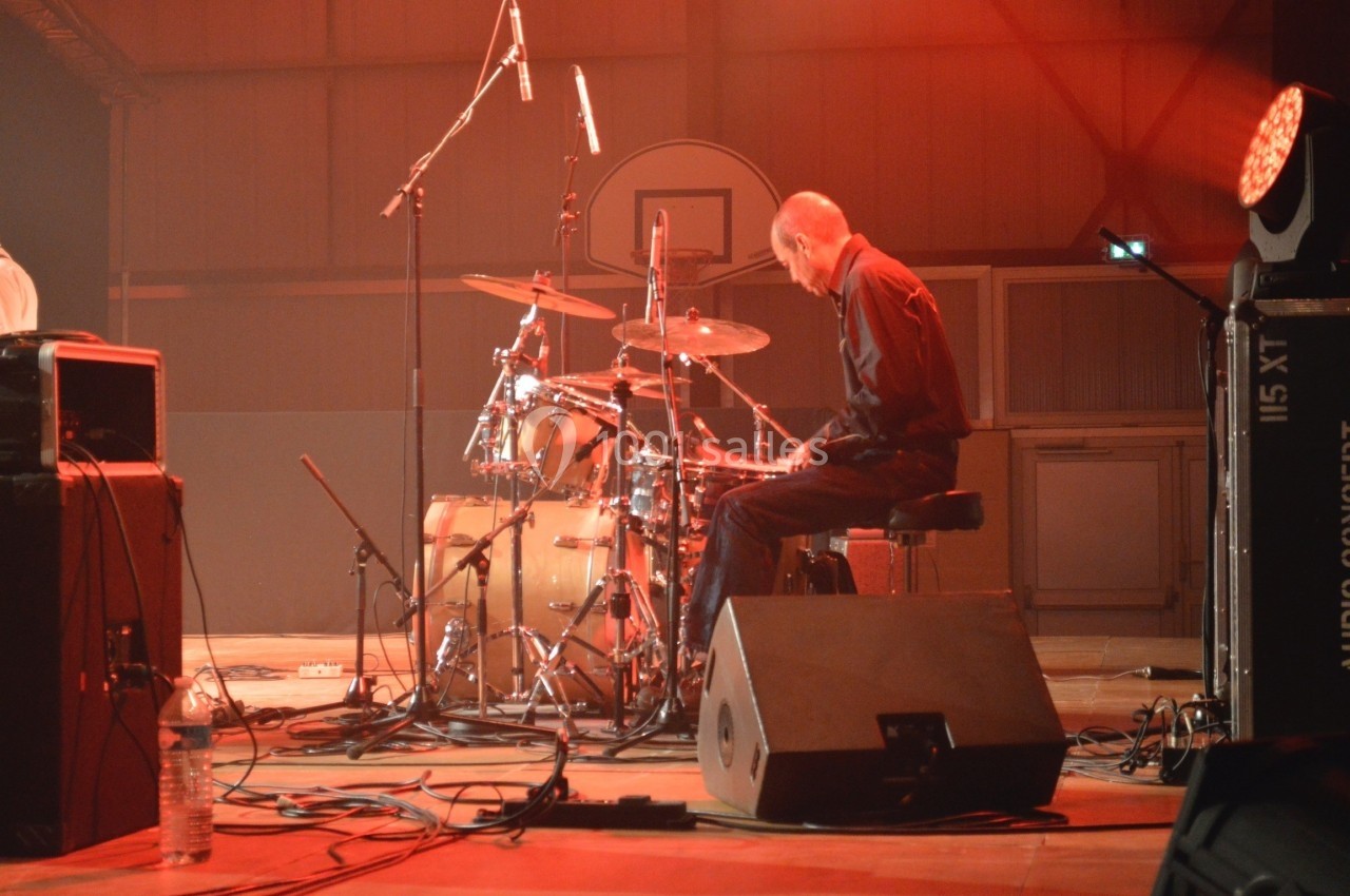Un batteur joue sur une batterie sur une scène éclairée de lumières rouges dans une salle de concert.