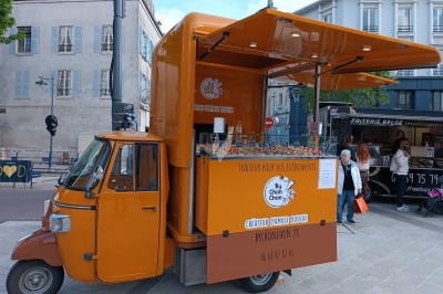 Triporteur orange aménagé en stand de traiteur, proposant des boissons et snacks sur une place urbaine.