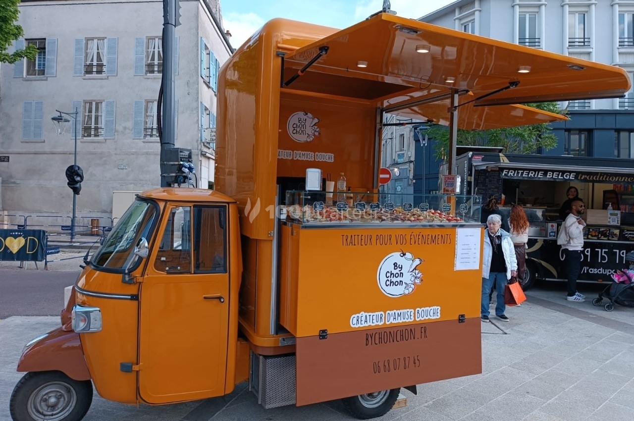 Triporteur orange aménagé en stand de traiteur, proposant des boissons et snacks sur une place urbaine.