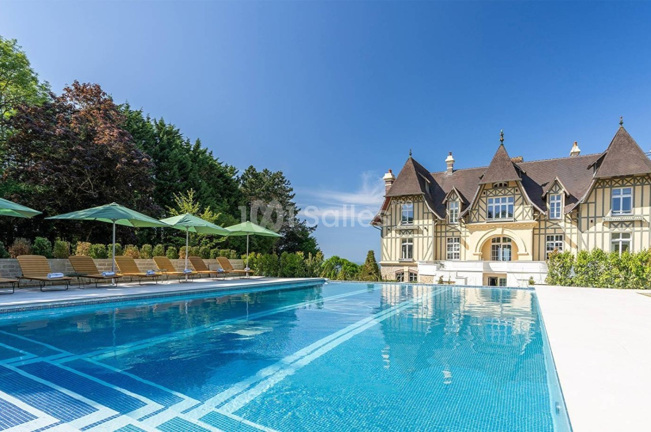 Piscine extérieure avec transats et parasols, devant un manoir de style traditionnel entouré de verdure.