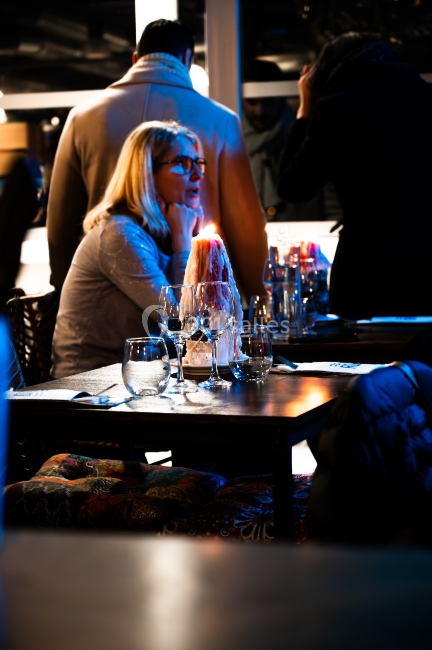 Une femme assise à une table de restaurant éclairée, entourée de verres et d'une bougie, avec des personnes en arrière-plan.