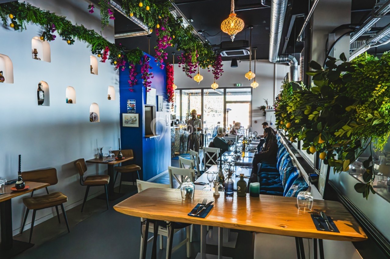 Salle de restaurant moderne avec décoration végétale suspendue, tables en bois et lumière naturelle provenant des baies…