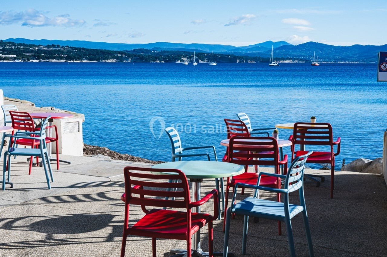 Chaises colorées et tables disposées en terrasse face à une mer calme avec des montagnes en arrière-plan.