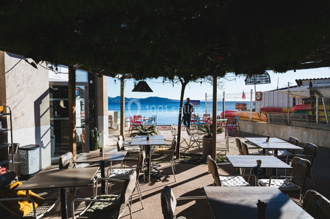 Terrasse ombragée d'un café avec vue sur la mer, tables et chaises disposées face au paysage côtier.