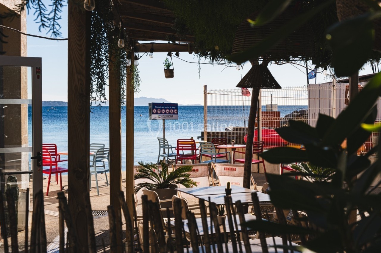 Terrasse ombragée avec vue sur la mer, mobilier coloré et végétation en premier plan.