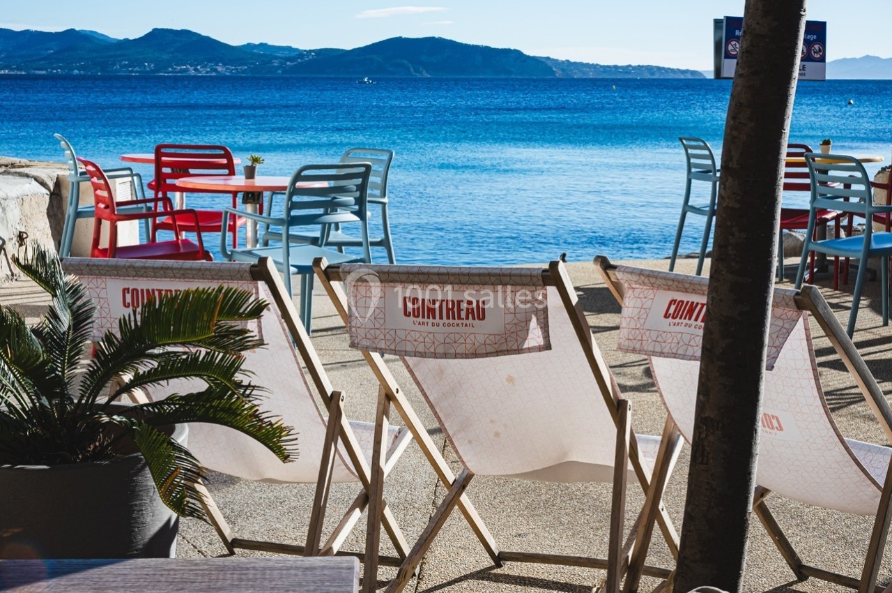 Chaises longues et tables colorées disposées face à la mer avec des montagnes en arrière-plan.