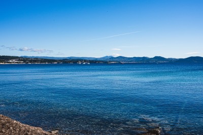 Vue d'un littoral avec une mer calme, un quai, des drapeaux et des collines à l'horizon sous un ciel dégagé.